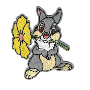 May include: Embroidered patch featuring a gray bunny holding a yellow flower with a green stem in its mouth. The bunny has tan accents and red eyes. The patch is on a white background.