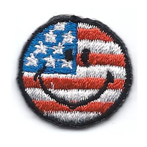 May include: A small, round embroidered patch with a smiley face design. The smiley face is made up of the American flag, with red and white stripes and blue stars.