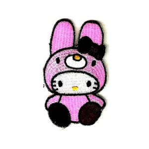 Op de afbeelding: Roze Hello Kitty-figuur met een roze konijnenkostuum met zwarte oren en een zwarte strik. Het figuur zit met gekruiste benen en heeft een gele neus en zwarte ogen.