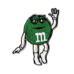 May include: Green M&M character with a white 'M' on its chest, wearing white gloves and boots, waving with its right hand.