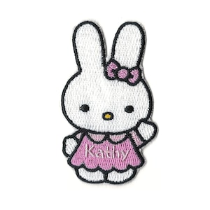 May include: White embroidered bunny with pink bow and dress. The dress has the name "Kathy" embroidered on it.