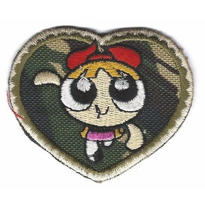 Może przedstawiać: Naszywka haftowana w kształcie serca z postacią z kreskówki. Postać to dziewczyna z blond włosami, czerwoną kokardką i dużymi oczami. Naszywka jest na tle kamuflażu.
