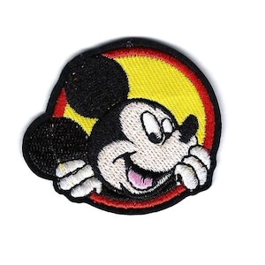 May include: Embroidered patch of Mickey Mouse. The patch has a black outline, a yellow and red background. Mickey Mouse is smiling with a pink tongue. The patch is circular in shape.