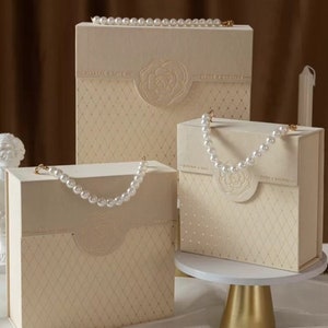 Puede incluir: Tres cajas de regalo de color marfil con asas de perlas. Las cajas est&aacute;n decoradas con un patr&oacute;n de diamantes y un dise&ntilde;o floral. La caja m&aacute;s grande est&aacute; a la izquierda, la caja m&aacute;s peque&ntilde;a est&aacute; a la derecha y la caja del medio est&aacute; en un soporte dorado.