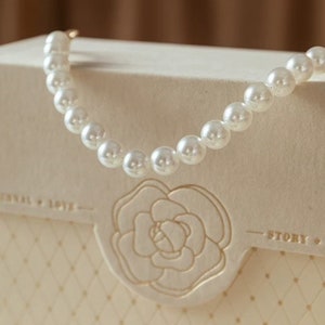 Puede incluir: Un collar de perlas blancas sobre una caja blanca con un dise&ntilde;o floral y el texto "Story".