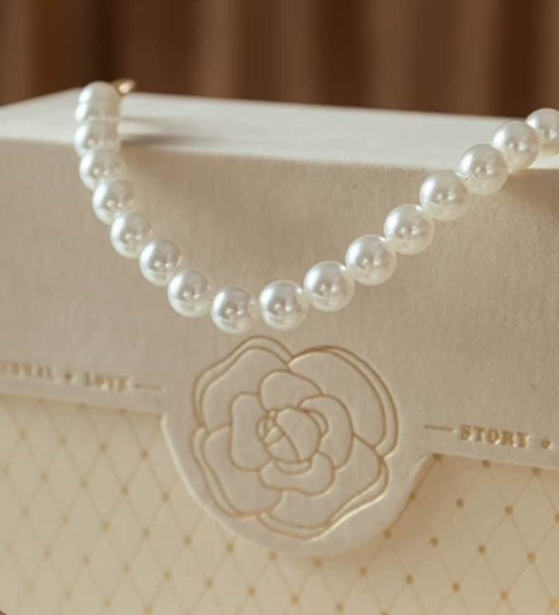 Puede incluir: Un collar de perlas blancas sobre una caja blanca con un dise&ntilde;o floral y el texto "Story".