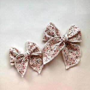 May include: Two hair bows of different sizes featuring a cream-colored fabric with a dense floral pattern in shades of red, green, and brown. Each bow has frayed, raw edges and a central knot detail, showcasing a soft, textured appearance.