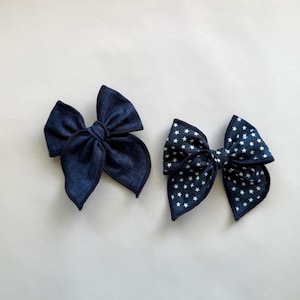 May include: Two dark blue fabric bows. One bow is solid, the other has a white star pattern. Both bows are tied in the center and have a layered design. The bows are likely hair accessories.