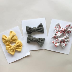 May include: Three pairs of fabric hair bows in various colors and patterns. One pair is yellow, another is gray, and the third is white with a red floral print. Each pair is displayed on a white card.
