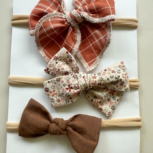 May include: Three headbands featuring fabric bows attached to thin, beige elastic bands. The top bow is rust-orange with a white grid pattern and frayed edges. The middle bow has a cream background with a small floral print. The bottom bow is solid brown.