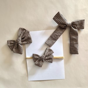 May include: Four brown and white gingham bows of varying sizes are arranged on a white surface. One bow is attached to a headband. The bows are made of fabric and have a classic, preppy style. The bows are suitable for hair accessories.
