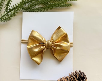 May include: A gold metallic bow on a beige headband, presented on a white card. The bow is the focal point, with a textured appearance. A sprig of green foliage and a pinecone are in the background, suggesting a festive theme.