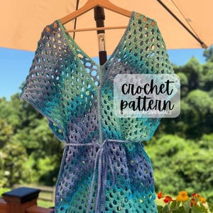 Op de afbeelding: Een handgemaakte, open geweven gehaakte cover-up in blauw- en groentinten, hangend aan een houten hanger. Het kledingstuk heeft een V-hals en een taille met trekkoord. De tekst "crochet pattern" is zichtbaar.