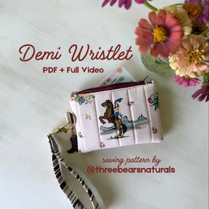 May include: A light pink quilted wristlet featuring a graphic of a person riding a horse, a zipper closure, and a black and white patterned wrist strap. The text reads Demi Wristlet, PDF + Full Video, and sewing pattern by @threebearsnaturals.