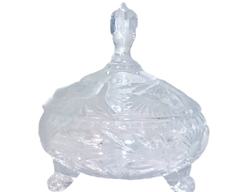 May include: A clear glass candy dish with a lid and three legs. The dish is decorated with a cut glass pattern.