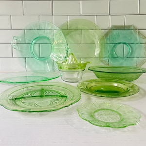 Può includere: Una collezione di oggetti in vetro pressato verde chiaro che include diversi piatti con motivi in rilievo, una ciotola poco profonda e uno spremiagrumi centrale. La cristalleria presenta vari design circolari, smerlati e divisi in tonalità di verde traslucido.