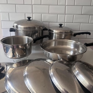 Könnte beinhalten: Ein Set aus Edelstahl-Kochgeschirr, bestehend aus einem großen Suppentopf mit Deckel, einem kleineren Stieltopf mit Deckel, einer breiten Pfanne und mehreren zusätzlichen runden Metalldeckeln. Jedes Teil hat schwarze Kunststoffgriffe.