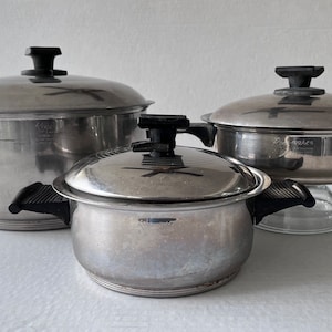 Può includere: Tre pentole in acciaio inossidabile con coperchi. Le pentole hanno manici e pomelli neri. I coperchi presentano un disegno a forma di croce. Le pentole sono di diverse dimensioni e poggiano su una superficie bianca. Le pentole sono etichettate con i marchi.