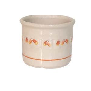 May include: A small, off-white ceramic container with a cylindrical shape. The container is decorated with orange and yellow candy corn designs around the middle and an orange stripe near the base. The rim is slightly flared.