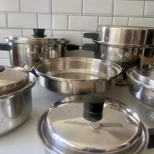 Op de afbeelding: Een verzameling roestvrijstalen kookgerei, waaronder potten, pannen en deksels. De potten hebben zwarte handvatten en knoppen. Het kookgerei staat op een wit oppervlak.