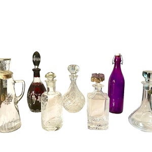 May include: A collection of seven glass decanters and bottles, including a purple glass bottle with a metal hinged lid, a clear glass decanter with a silver top, and a red glass decanter with a black stopper.