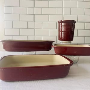 Può includere: Set di quattro teglie da forno in ceramica rossa. Le teglie sono di diverse dimensioni e forme, con un interno color crema.