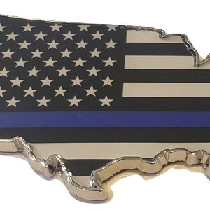 May include: A metal outline of the United States of America with a thin blue line across the middle of the American flag design. The flag is black, white, and blue.