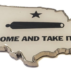 May include: A white metal outline of the United States with a black silhouette of a cannon and a single star on a white background. The text "COME AND TAKE IT" is printed below the cannon.