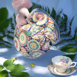 Peut inclure: Une théière recouverte d'un motif de plaques colorées est tenue au-dessus d'une tasse et d'une soucoupe. La tasse a un motif floral avec des bordures dorées. Des sachets de thé sont sur la surface bleu clair. Des fougères et des feuilles sont en arrière-plan.