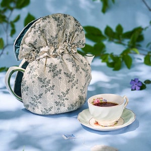 May include: A cream-coloured teapot with a floral tea cosy and a teacup on a saucer. The cosy has a green lining and a pattern of small, dark green leaves. The teacup is filled with tea and has a floral design.