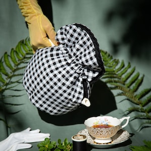 May include: A black and white gingham tea cosy covering a teapot, positioned above a floral teacup and saucer filled with tea, accompanied by a spool of black thread and white gloves, all set against a muted green backdrop with fern leaves.