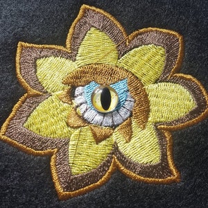 May include: Embroidered flower patch with a brown and yellow design. The center of the flower has a large, blue eye with a yellow iris and a black pupil.