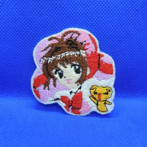 May include: Embroidered patch featuring a cartoon girl with brown hair and a red outfit, with a small yellow bear character. The patch has a pink and white border and is set against a blue background.
