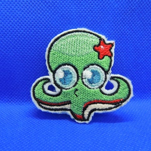 Peut inclure: Pieuvre brodée verte avec de grands yeux bleus et une étoile rouge sur le dessus. La pieuvre est bordée de blanc et a une bouche rouge.