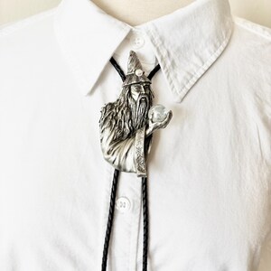 May include: A bolo tie featuring a silver-toned metal pendant of a bearded wizard wearing a pointed hat and robes, holding a small clear sphere in one hand. The pendant is attached to a black braided cord worn over a crisp white collared shirt.