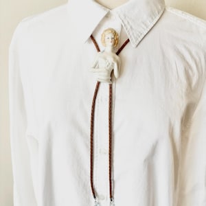 May include: A bolo tie featuring a porcelain half-doll bust with blonde hair and a white bodice. The tie consists of two braided brown leather cords that hang down from the central figure, each finished with a silver-toned metal tip and blue beads.