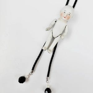 May include: A bolo tie featuring a small, white porcelain doll figure with painted facial features. The doll is attached to a black braided cord that ends in silver-toned metal caps, each accented with a small white bead and a faceted black bead.