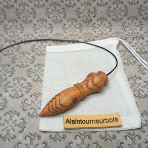 Peut inclure: Un pendule en bois avec une extrémité pointue et un motif en chevrons sculpté. Le pendule est attaché à un cordon noir et repose sur un petit sac en tissu blanc cassé. Le sac est posé sur un tissu à motifs. L'étiquette en bois indique "Alaintournebois".