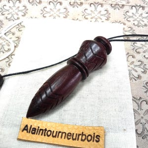 Peut inclure: Un pendule en bois violet foncé avec une pointe effilée et des motifs en chevrons sculptés, attaché à un cordon noir fin. Une étiquette rectangulaire en cuir beige avec le texte noir Alaintourneurbois est placée sous l'objet en bois.