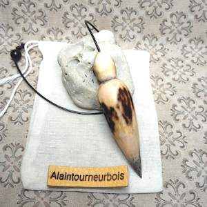 May include: A wooden pendulum with a teardrop shape and dark brown grain patterns, attached to a black cord. The pendulum rests on a light-colored stone and a small white fabric pouch featuring a tan leather tag with the text Alaintourneurbois.