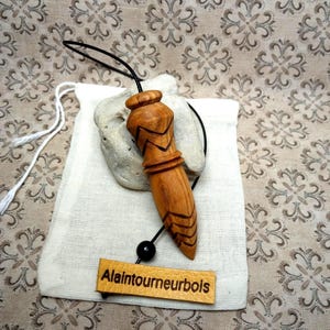 Puede incluir: Un péndulo de madera de color marrón claro con patrones de chevrón tallados, unido a un cordón negro con una pequeña cuenta negra y una etiqueta rectangular de cuero beige con el texto Alaintourneurbois, sobre una bolsa de tela blanca.