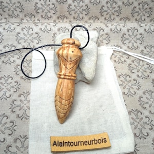 Puede incluir: Un péndulo de madera de color marrón claro con patrones geométricos tallados y un cordón negro. El péndulo descansa sobre una bolsa de tela blanca con una etiqueta de cuero beige con el texto Alaintourneurbois impreso en negro sobre un fondo estampado.