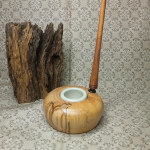 Puede incluir: Un portaplumas de madera con un inserto de cerámica blanca y una pluma de madera. El portaplumas está hecho de madera clara con vetas marrones oscuras. La pluma es larga y delgada con un acabado marrón oscuro.