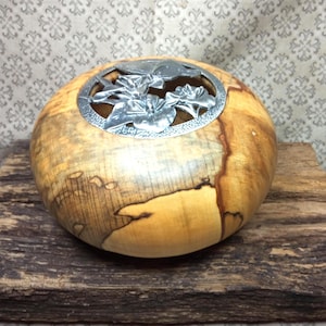 Peut inclure: Un bol en bois poli et arrondi présentant un grain naturel beige clair avec des stries et des nœuds brun foncé. L'ouverture supérieure est dotée d'un insert circulaire en métal argenté orné d'un motif floral en relief de feuilles et de pétales.