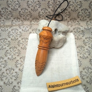 Könnte beinhalten: Ein geschnitztes Holzpendel mit spitzer Form und geometrischen Mustern, befestigt an einer schwarzen Schnur. Es liegt auf einem weißen Stoffbeutel neben einem hellen Stein. Ein kleines Holzschild mit dem Text Alaintourneurbois liegt bei.