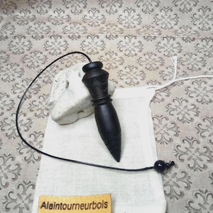 Puede incluir: Un péndulo de madera marrón oscuro con punta afilada y detalles geométricos tallados, unido a un cordón negro. Descansa sobre una bolsa de tela blanca con una etiqueta rectangular color canela con el texto impreso Alaintourneurbois.