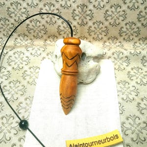 May include: A light brown wooden pendulum with carved chevron patterns, attached to a black cord with a single black bead. A small yellow tag with the text Alaintourneurbois is placed on a white fabric pouch behind the pendulum.