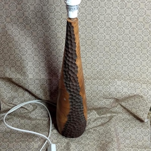 Peut inclure: Lampe de table en bois avec un motif sculpté. La lampe a une forme conique avec une finition marron clair et une section sculptée marron plus foncée. Elle possède un support de lampe blanc et un cordon blanc avec un interrupteur.