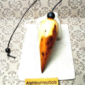 May include: A polished wooden pendulum with a teardrop shape, featuring natural light and dark brown wood grain patterns. It is attached to a black cord with a small black bead. A yellow leather tag with the text Alaintourneurbois is included.