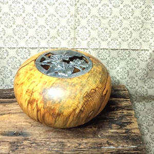 Peut inclure: Un bol en bois poli de forme ronde présentant un grain brun clair naturel avec des stries sombres. L'ouverture supérieure est recouverte d'un insert métallique argenté circulaire orné d'un motif floral découpé de feuilles et de fleurs.